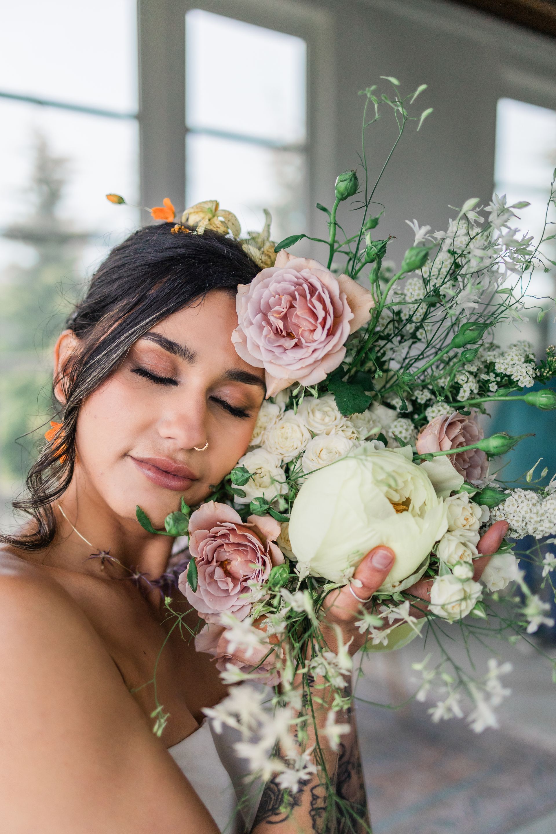 Woman holding a large pastel floral bouquet, eyes closed, near a sunlit window.