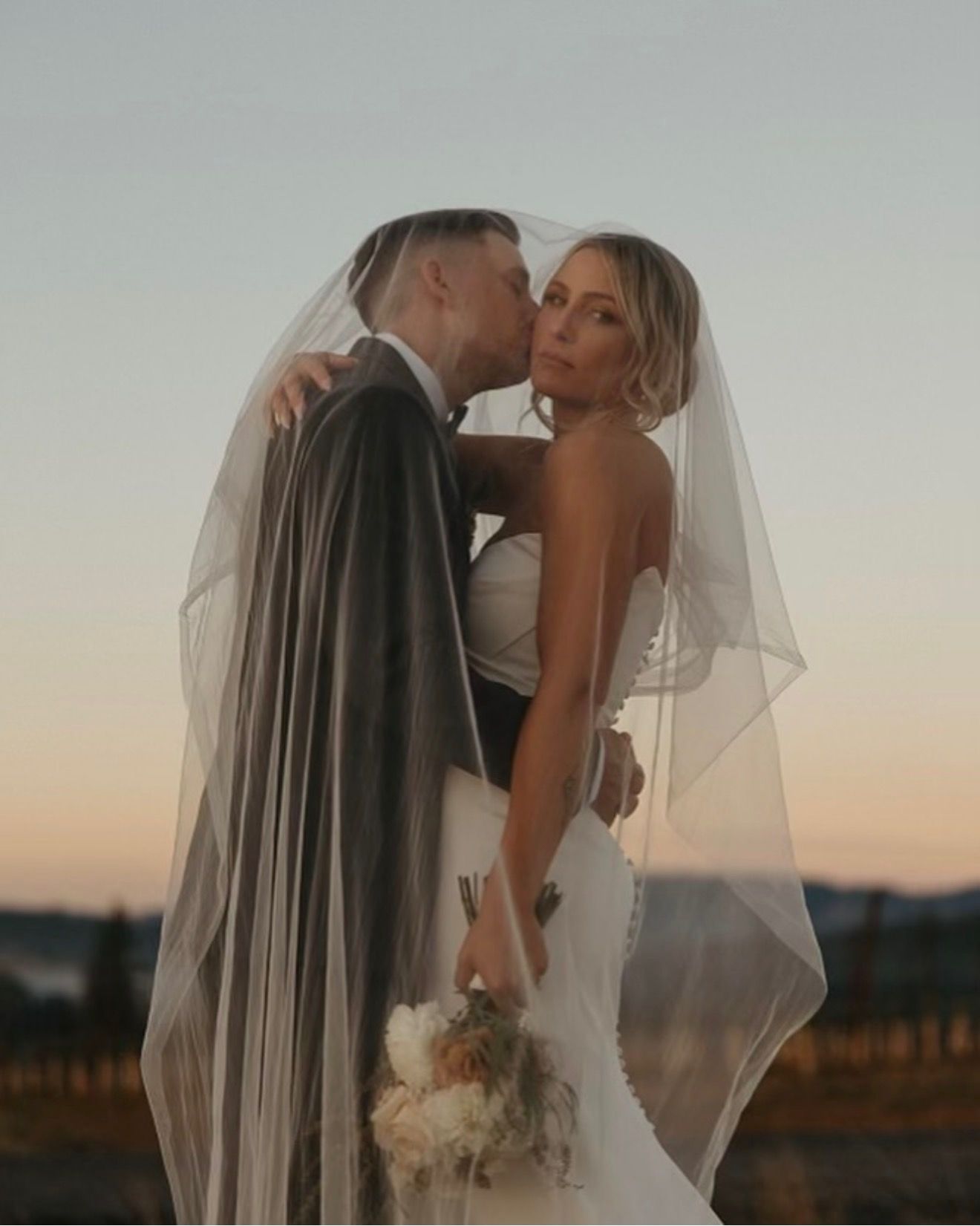 Bride and groom kiss at sunset, bride in white gown and veil holding bouquet.
