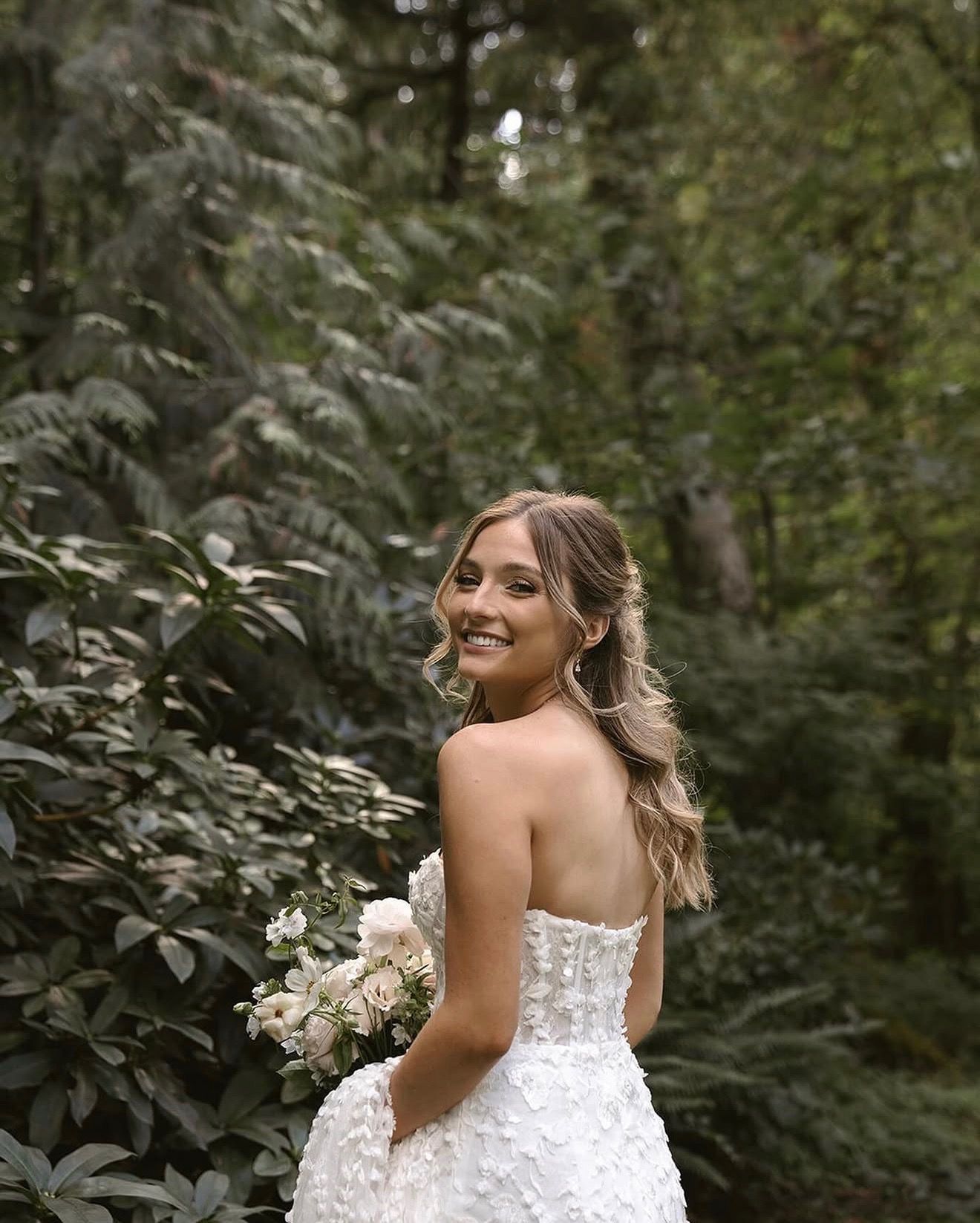Bride in a white dress smiling in a forest, holding a bouquet of white flowers.
