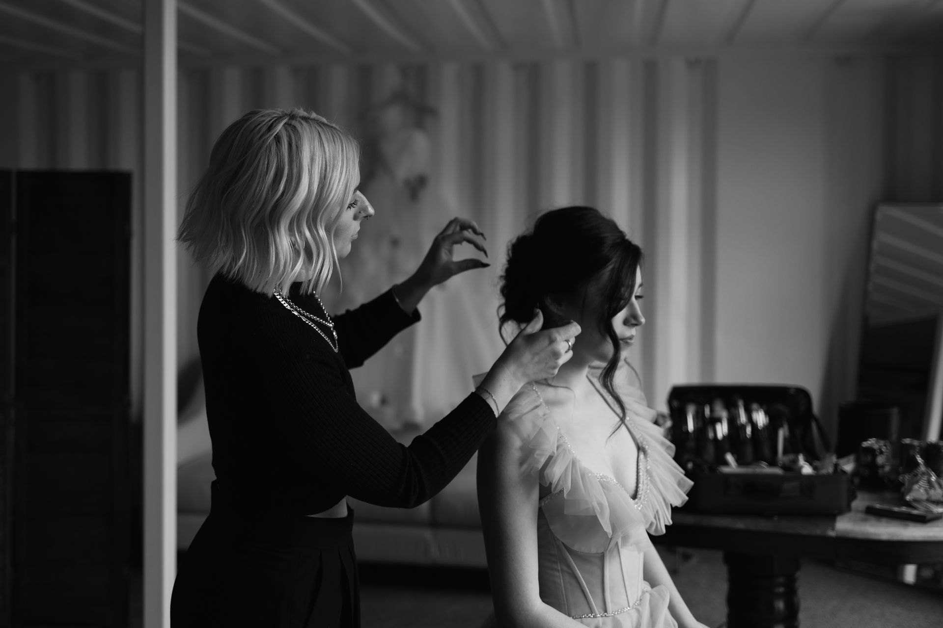 Hair stylist adjusting a bride’s updo in a softly lit room with a mirror and styling tools