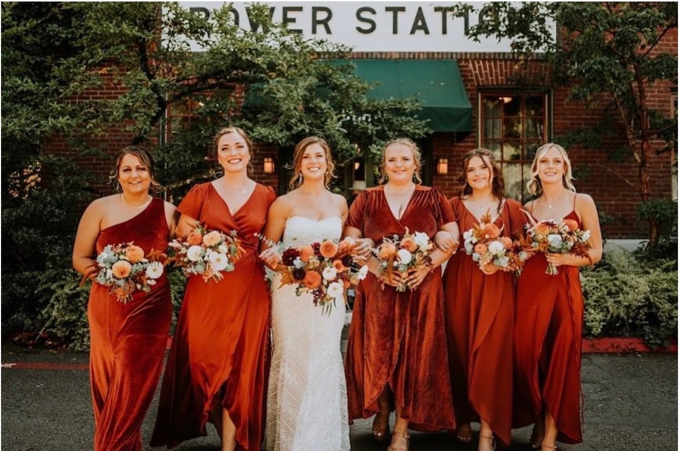 Six women in rust dresses holding bouquets stand in front of a flower-covered building entrance.