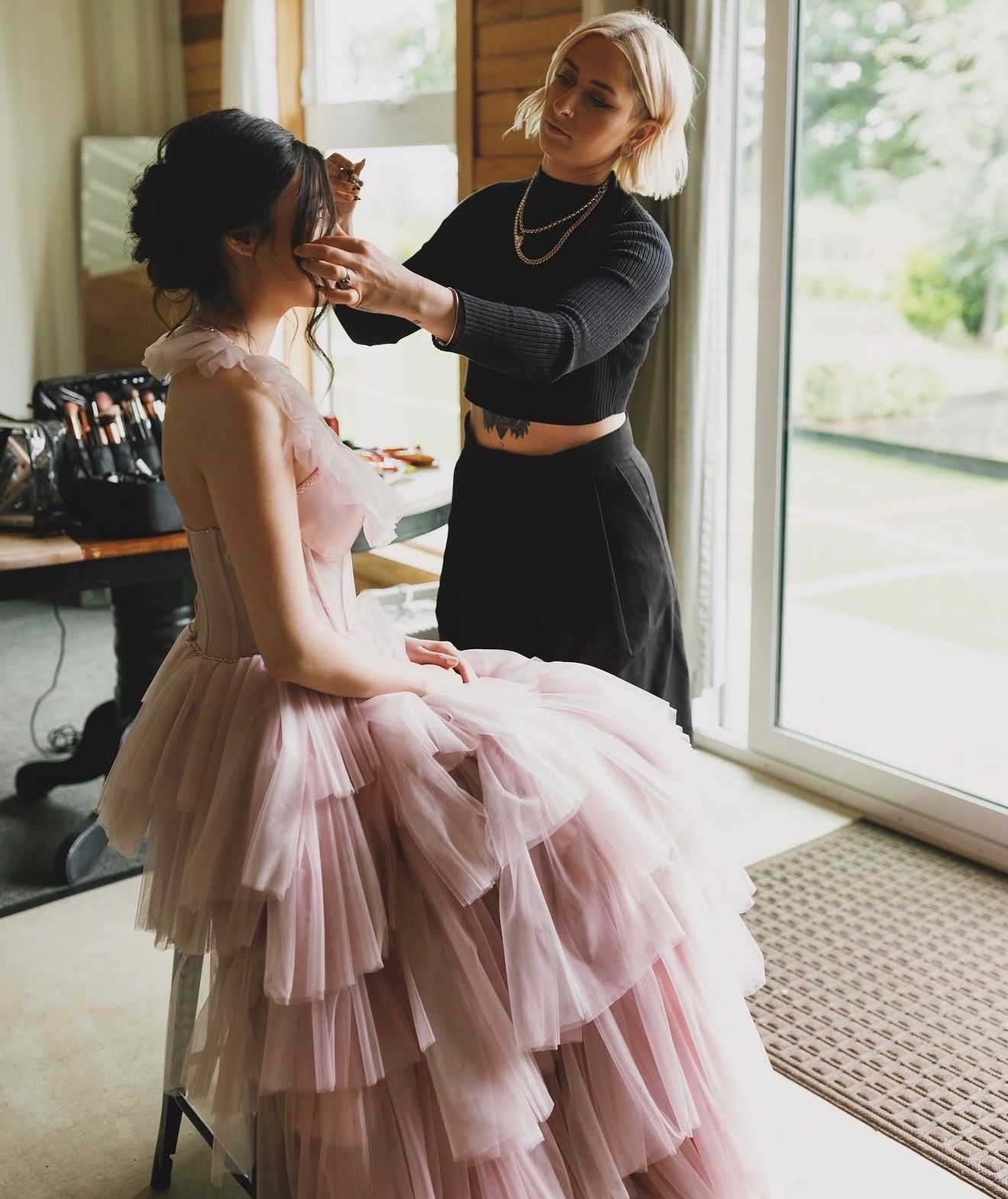 Makeup artist applying makeup to a seated woman in a pink tulle dress near a window