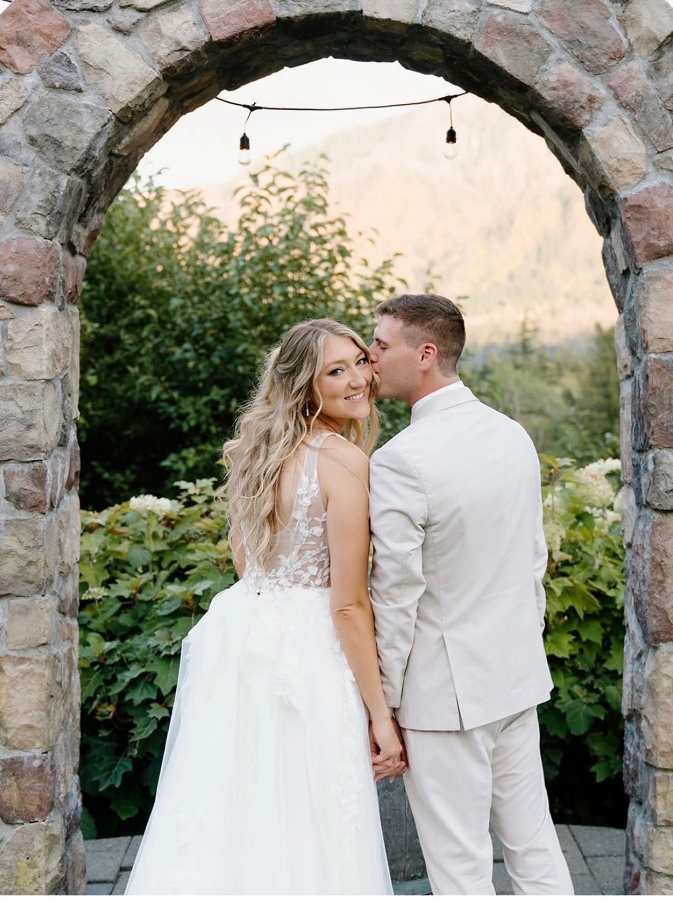 Couple holding hands beneath a stone arch in a garden, with the bride in a white gown and groom in a light suit