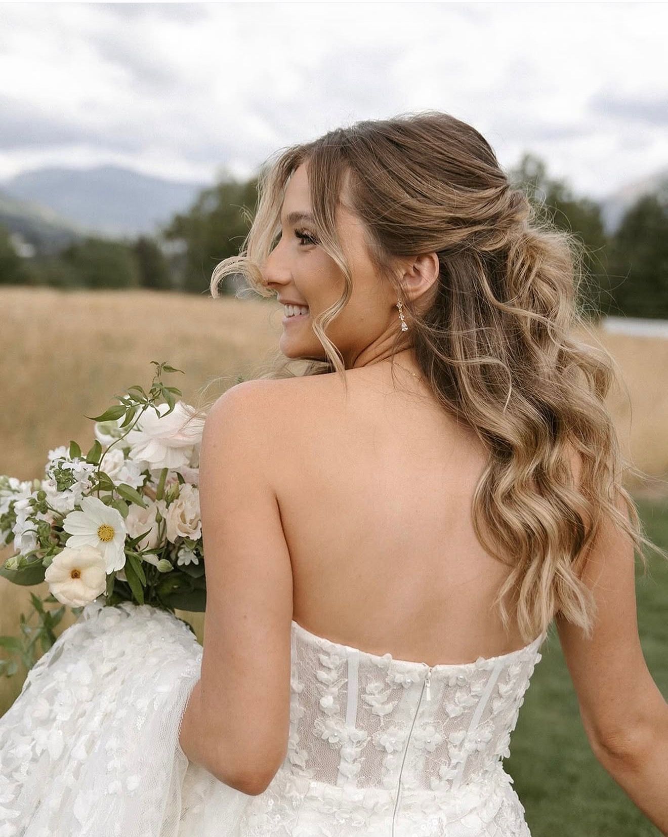 Bride in a white lace dress holding a bouquet, looking over her shoulder in a field with mountains behind her