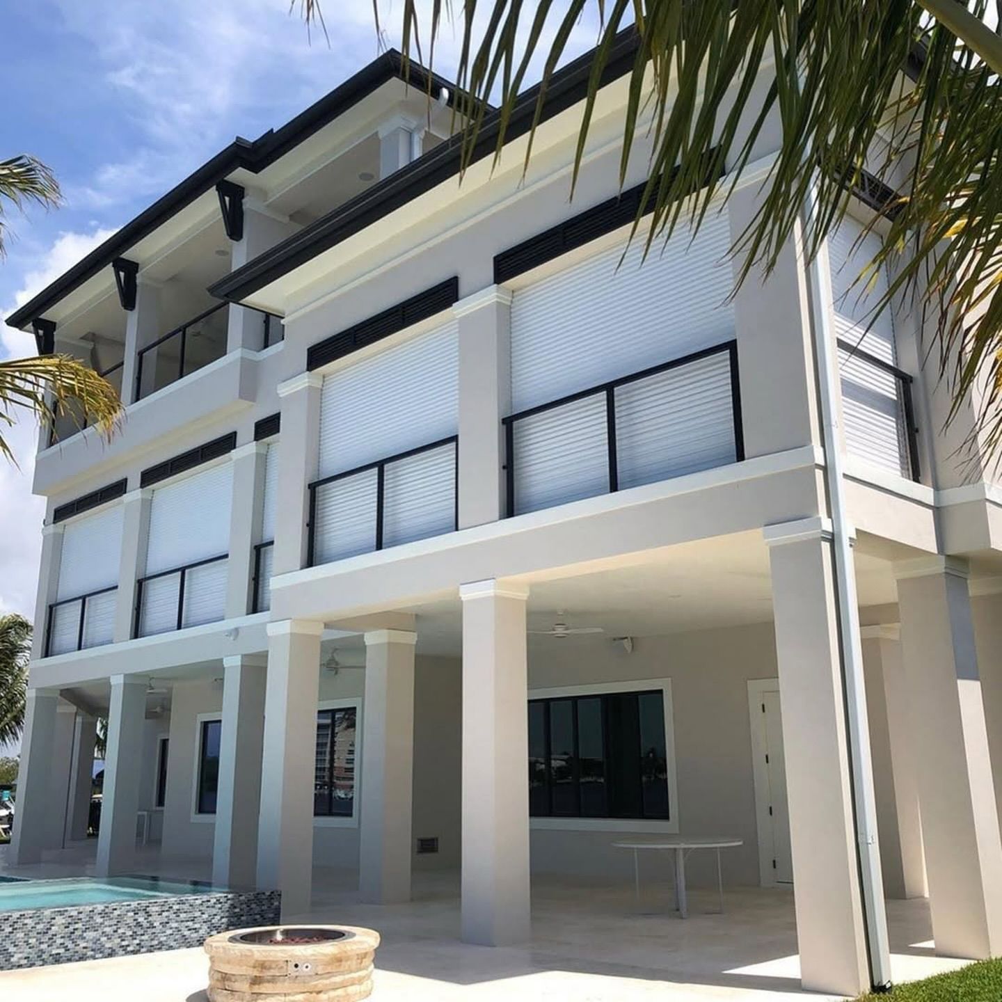 Three-story beige house with roll-down shutters, balconies, and black railings. Palm trees and a pool are visible.
