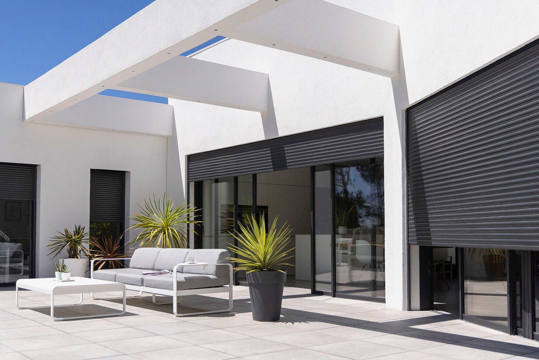 White patio with modern outdoor furniture, partially shaded with a dark roller shutter, and large windows.