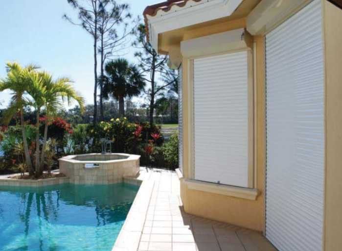 White roll-down shutters on a light yellow house next to a swimming pool and tropical landscaping on a sunny day.