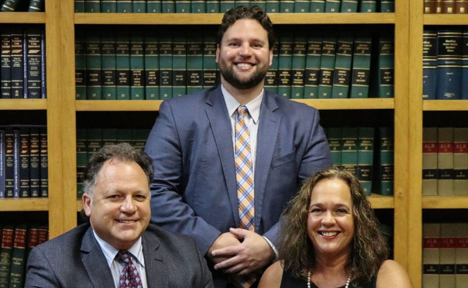 Three people in suits smiling in front of bookshelves in a library or law office.