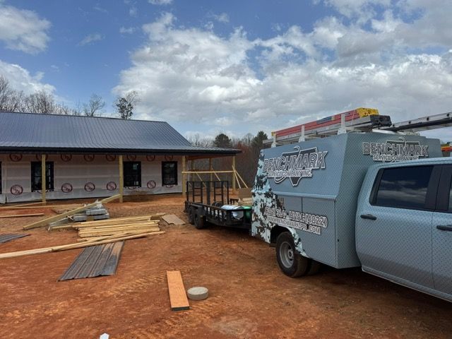 A partially constructed house with a trailer and truck in a dirt lot under a cloudy sky.