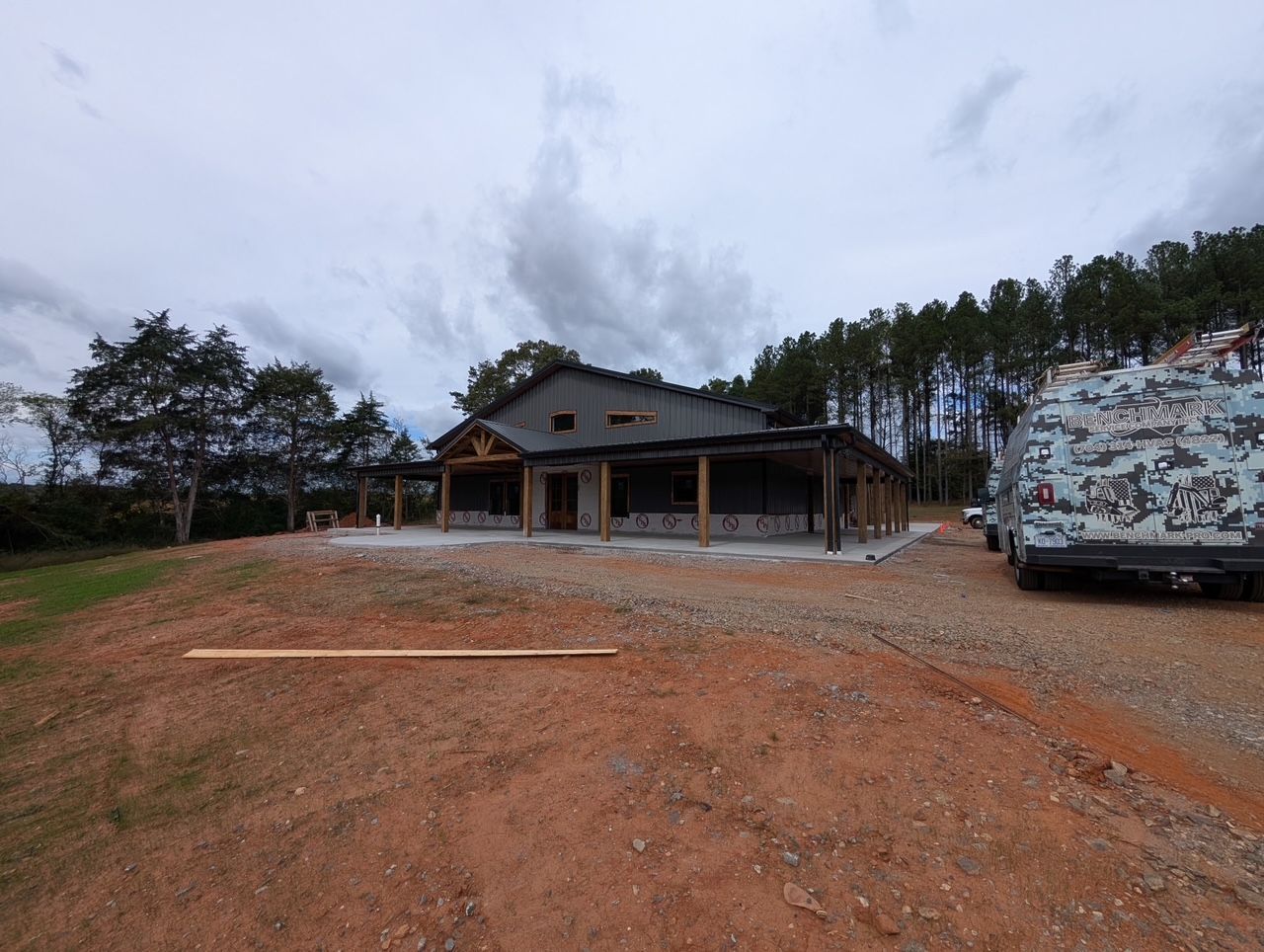 A partially constructed building with a porch under an overcast sky. A truck is parked nearby on a dirt lot.
