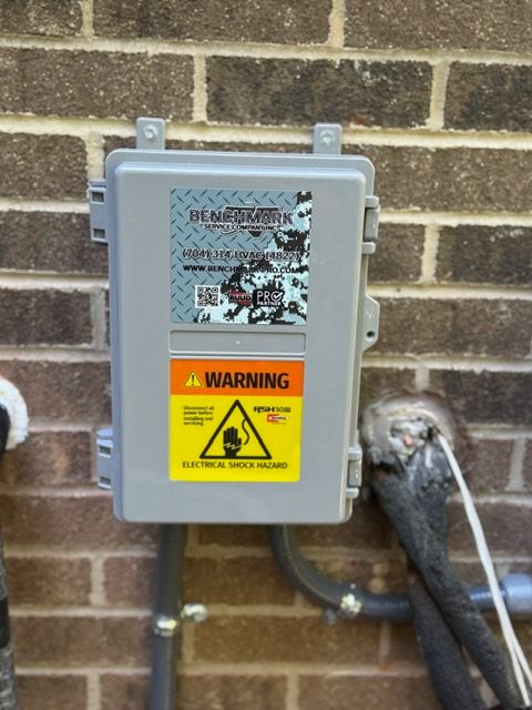 Gray electrical box on brick wall, with warning labels. Black conduit and pipe attached.