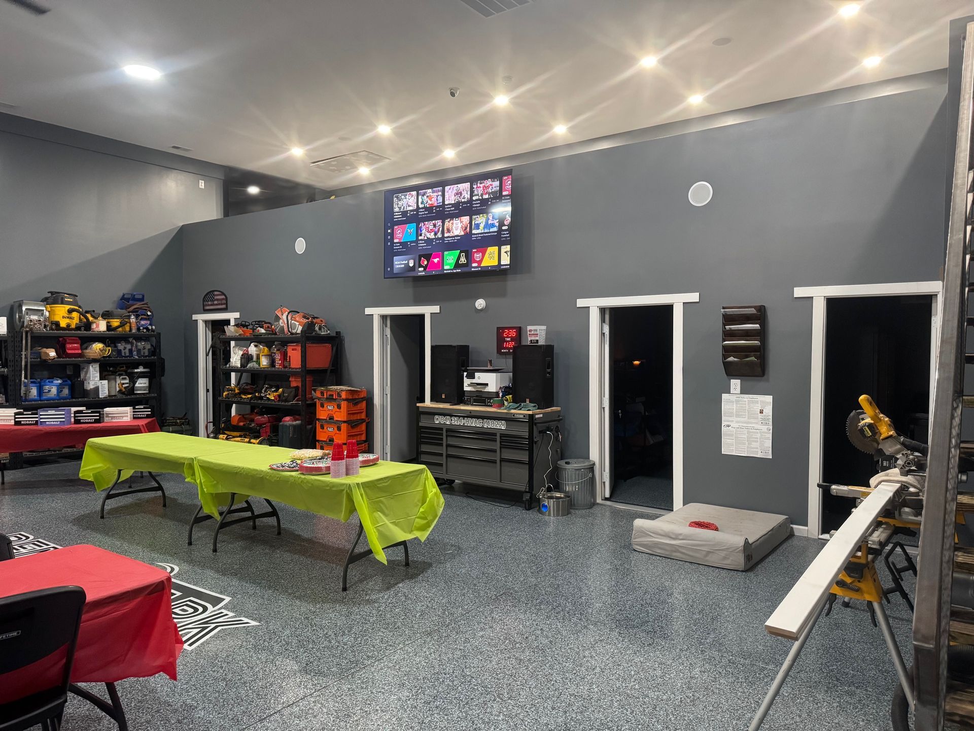 Workshop space with tables, tools, TV screen, and doors. The walls are gray, and the floor has a speckled blue pattern.