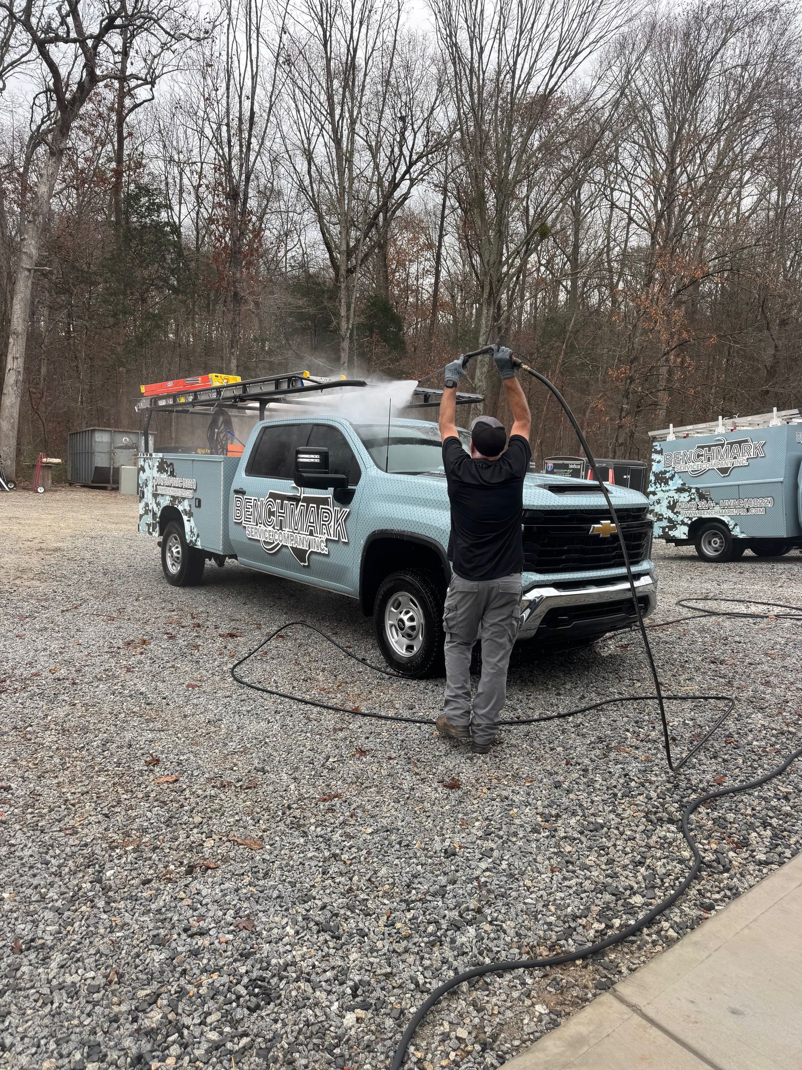 Person power washing a light blue truck with a logo parked on a gravel lot; another smaller trailer is visible.