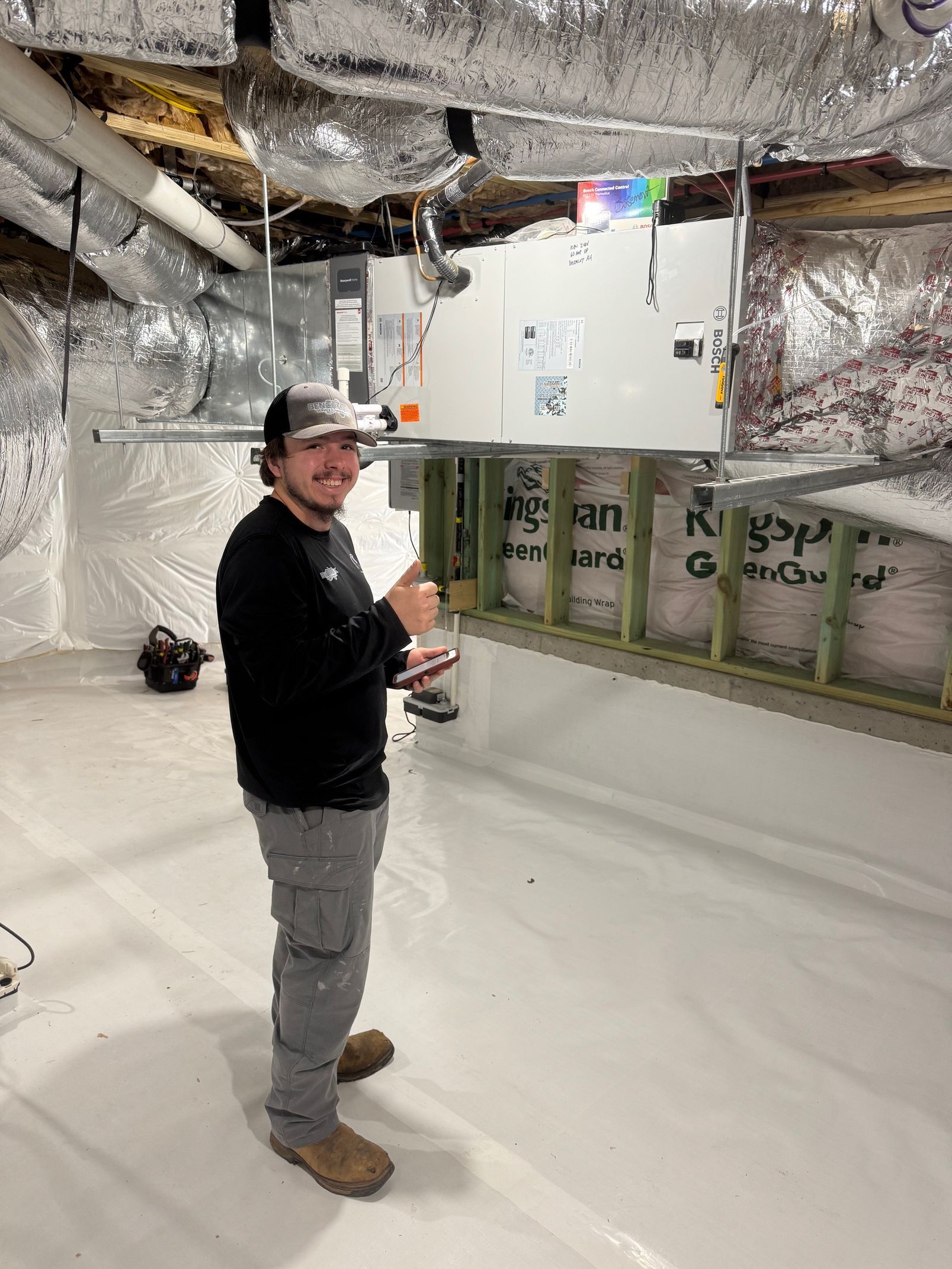 Man in work clothes standing in a crawl space, smiling, near HVAC equipment. White walls and insulation.
