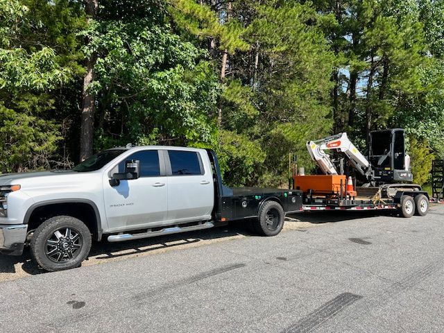 Silver truck towing a trailer with a small excavator on a paved road next to trees.