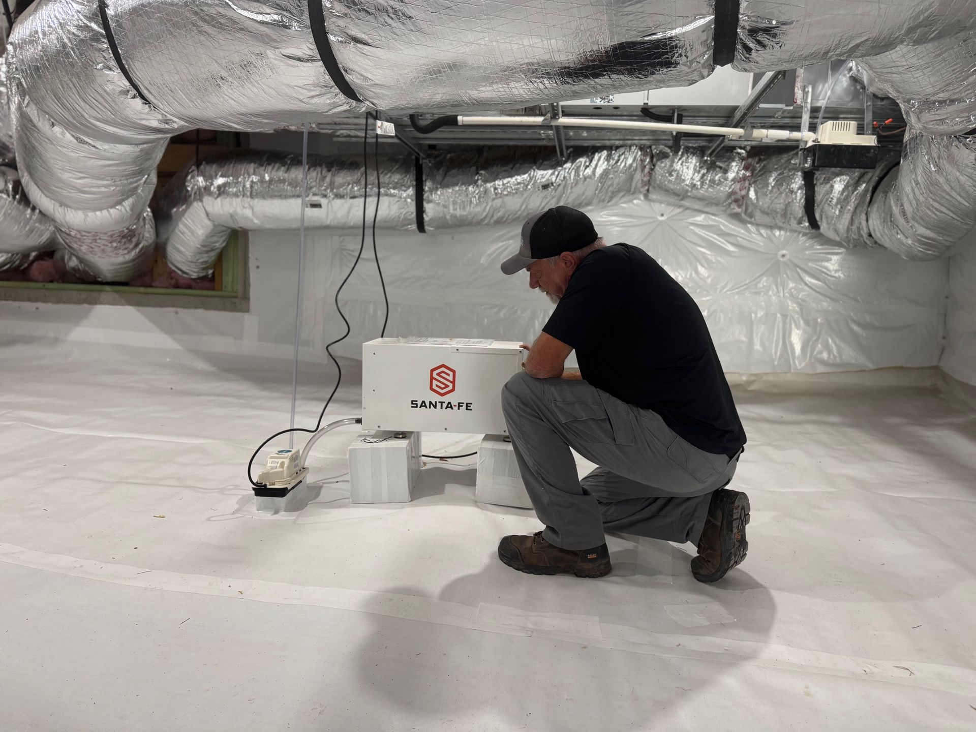 Man inspecting a white box unit in a crawl space with insulated ducts and white walls.