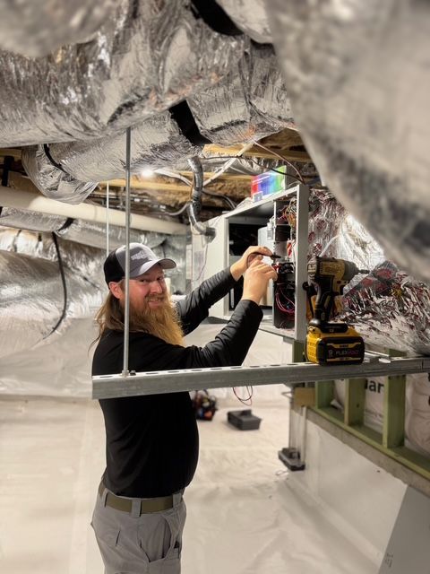 A man with a beard works on electrical components within a crawl space, using a drill.
