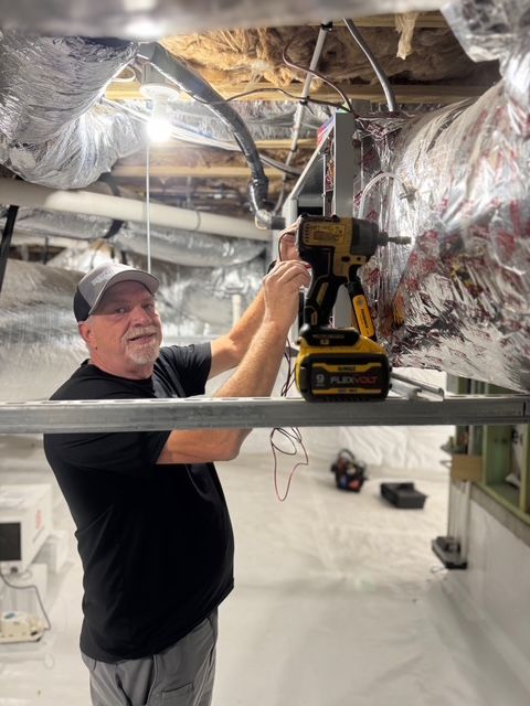 Man using a power drill on a metal frame, inside a crawl space with insulation and ductwork.