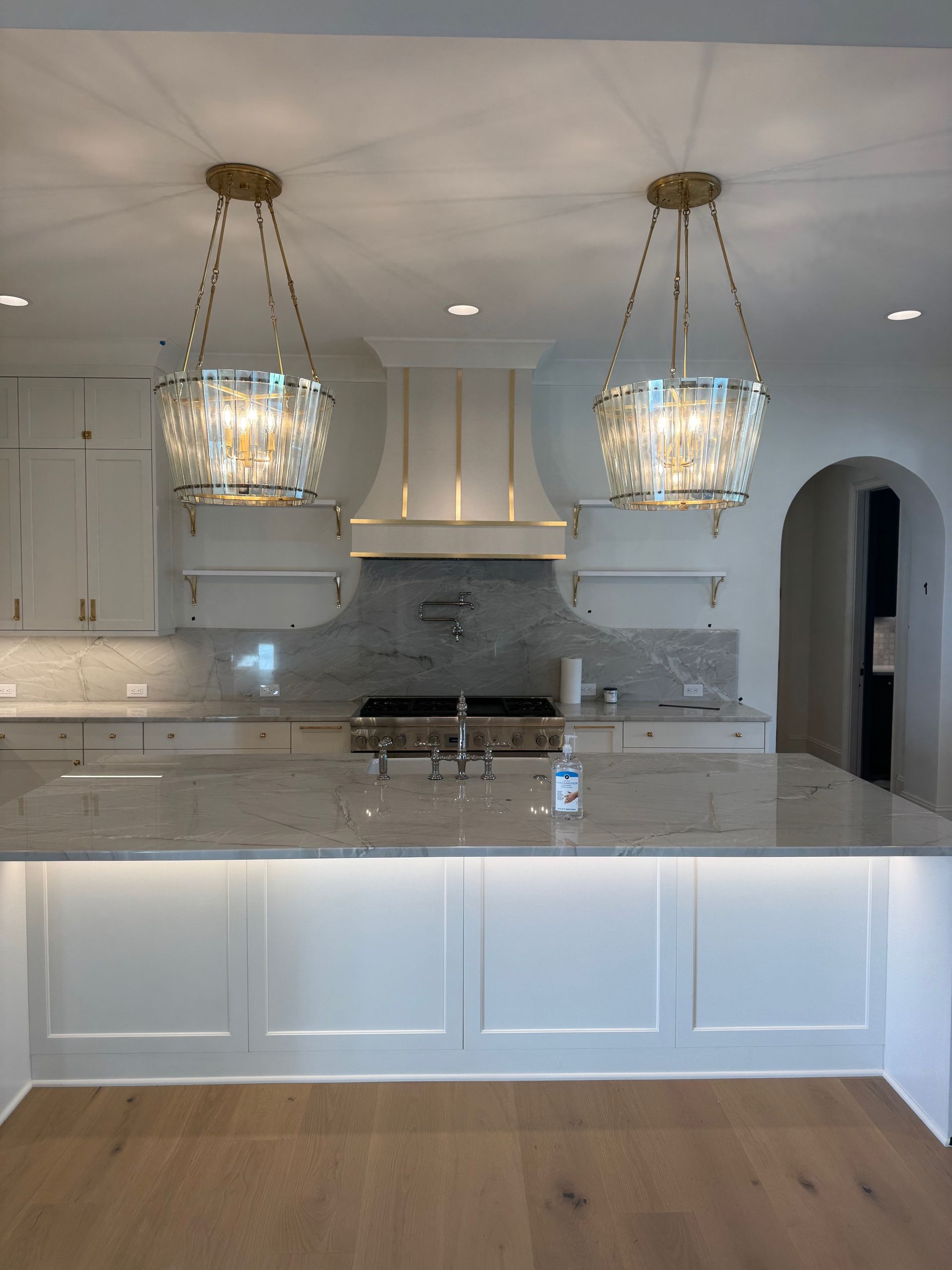 Kitchen with two crystal pendant lights, white cabinetry, marble countertops, and a range hood.
