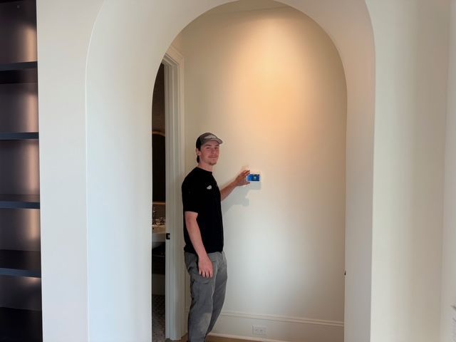 Man holding a small blue object, standing in an arched doorway of a home. White walls and a dark doorway are visible.