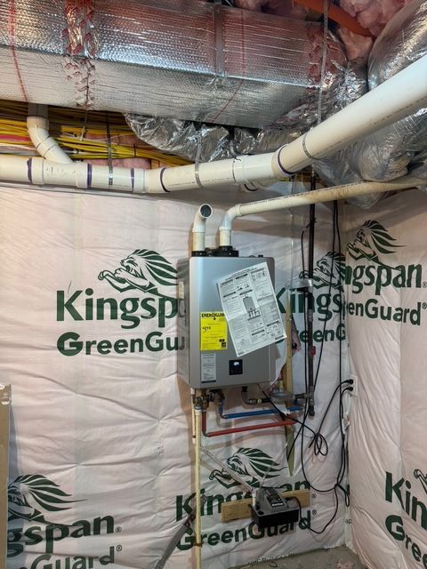 A wall-mounted, tankless water heater in a basement. Pipes and wires are visible, surrounded by insulation.