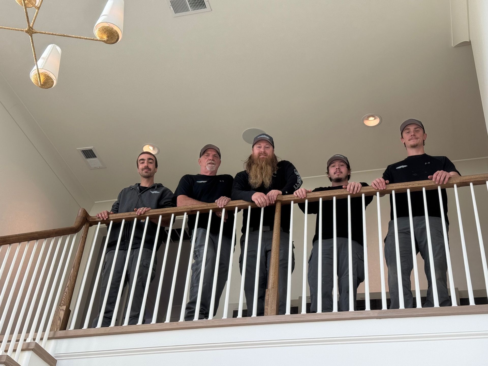 Five men stand on a balcony, smiling. They wear hats, black shirts, and grey pants. Wooden railing. Overhead chandelier.