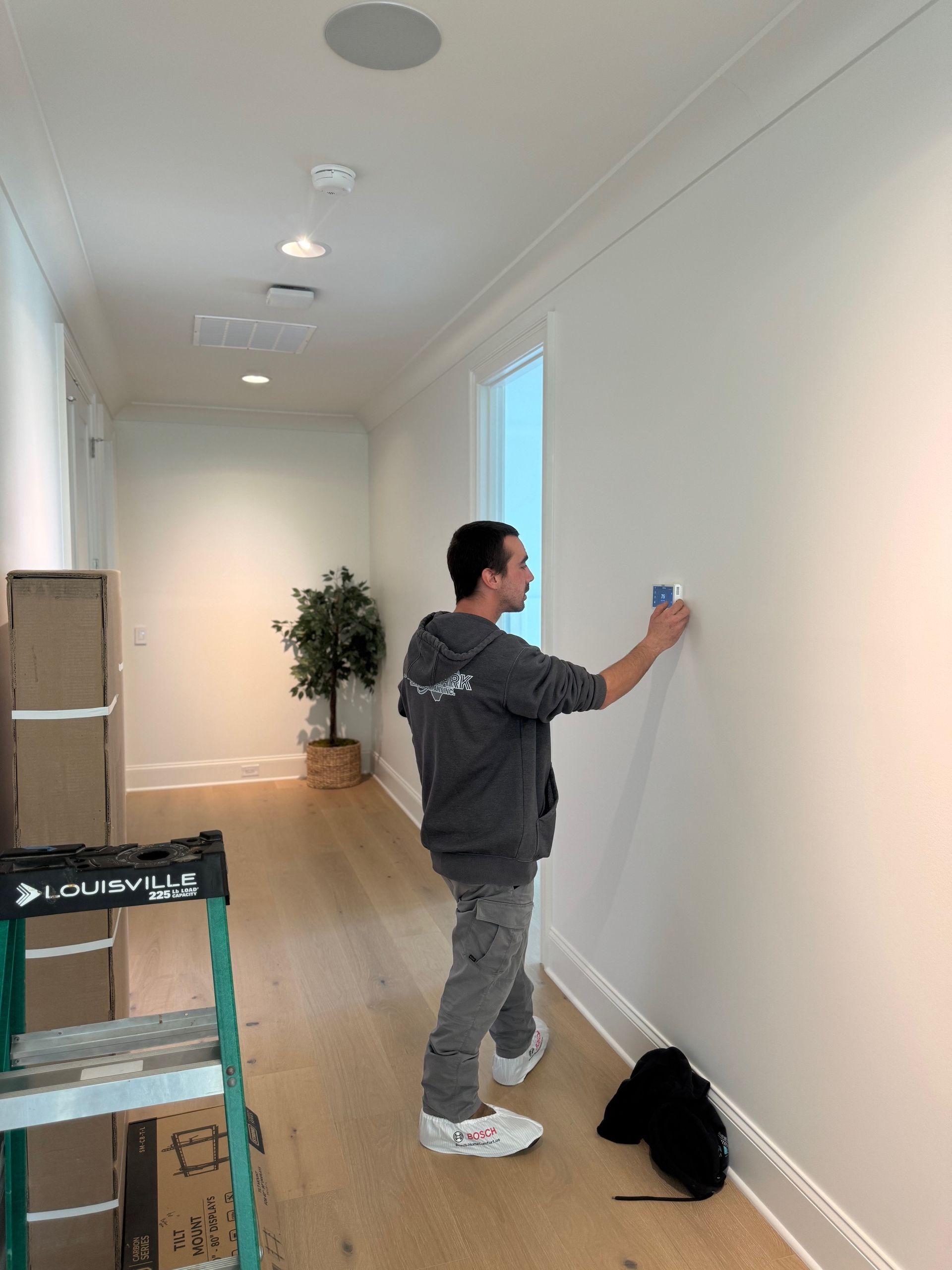 Man in gray hoodie installing something on a white wall in a hallway with wood floors.