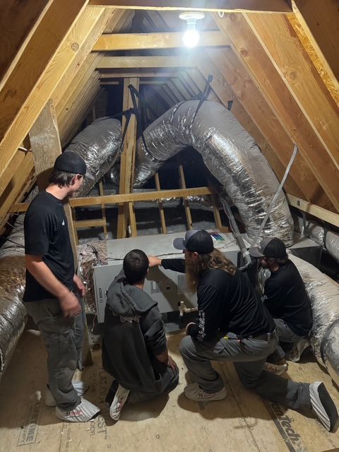 Four people installing HVAC equipment in an attic. They are near ducts and wooden beams.