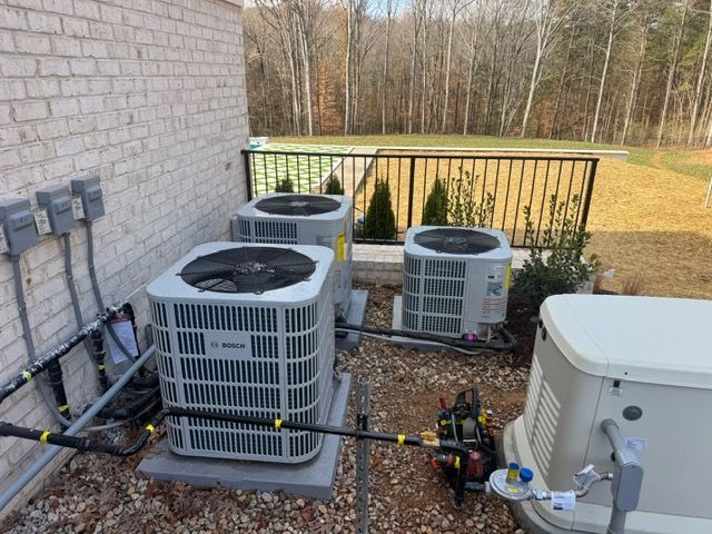 Three HVAC units and a generator next to a brick wall. Black pipes and wiring. Stone, gravel, and grass surround.