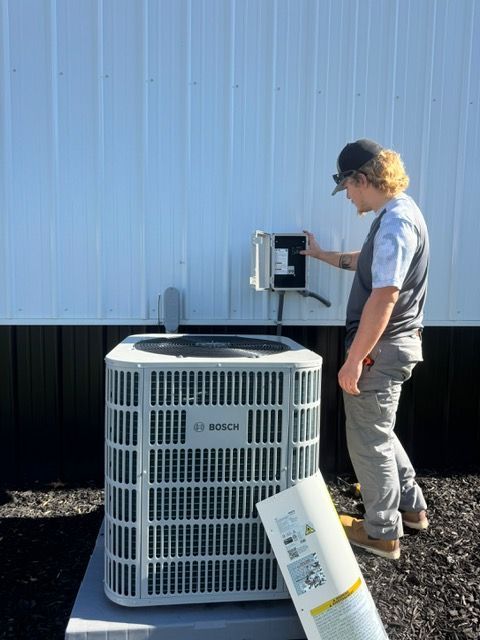 Person examining electrical box above Bosch air conditioner. Gray siding and concrete base.