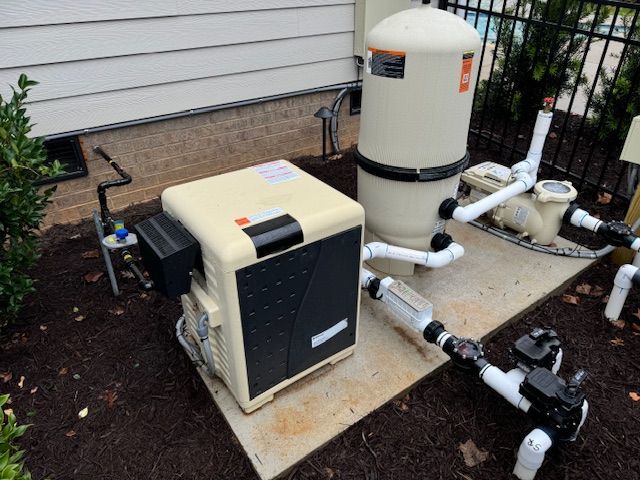 Pool equipment: heater, filter, and pump, on concrete pad, near house and fence, on mulch.