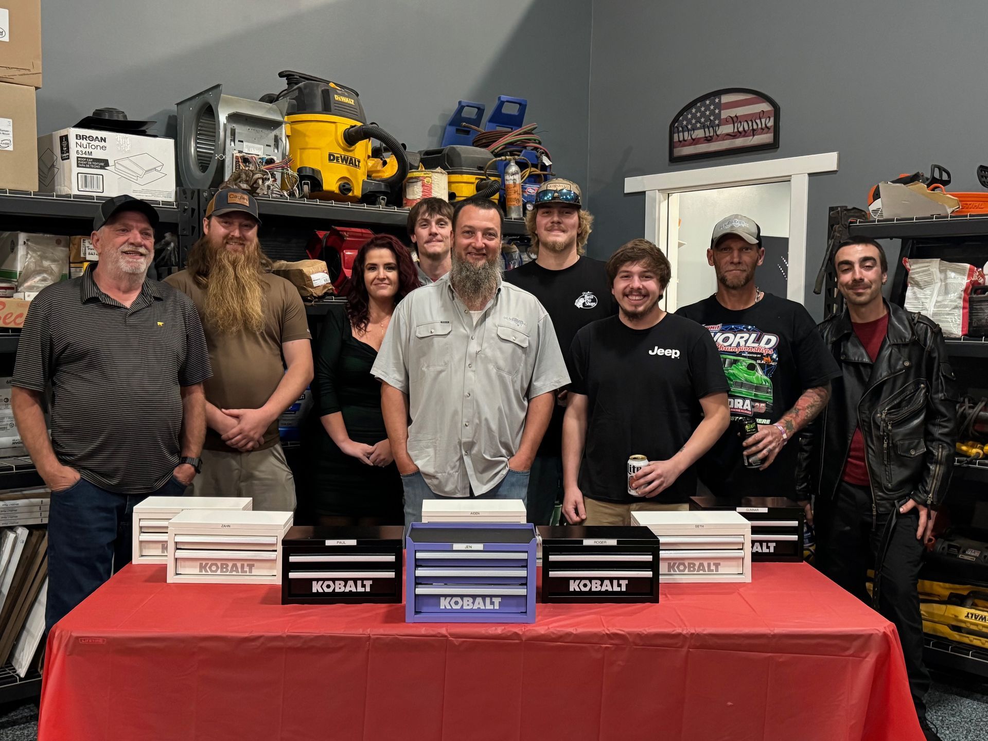 Group of people pose behind toolboxes on a red table in a shop setting. Tools on shelves in background.
