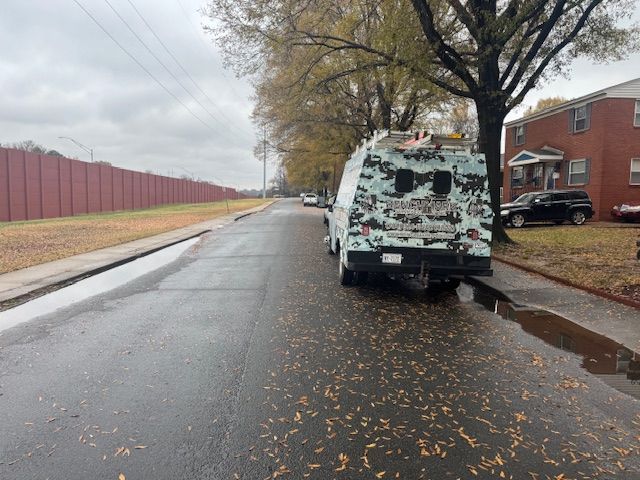 Wet street with a parked camouflage van, brown fence and brick buildings under a cloudy sky.