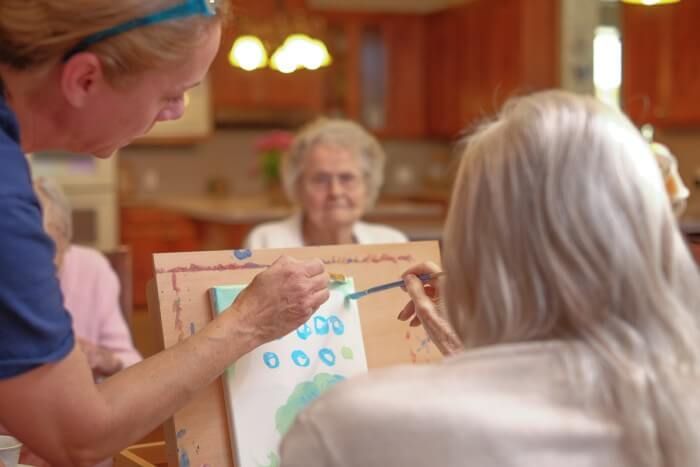 Woman helps another paint at a table; another woman looks on in a sunny room.