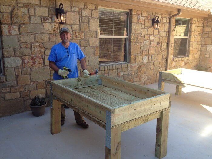 Man with a drill stands near a wooden raised garden bed on a concrete patio, stone wall in background.