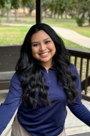 Woman with long dark hair smiles, wearing a navy blue shirt, standing outdoors near a wooden structure.