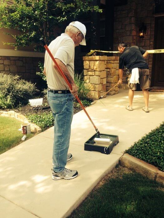 Two people painting a sidewalk. One uses a roller, the other brushes. House in background.