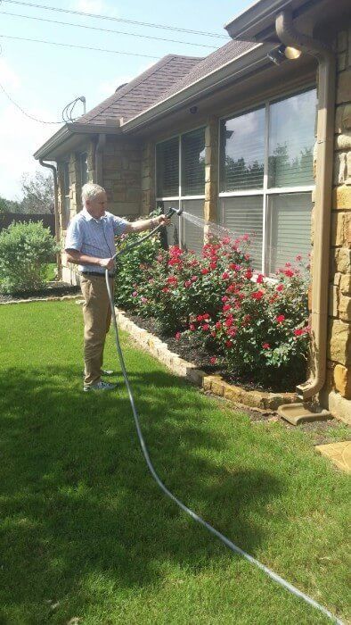 Man watering red rose bushes next to a house with a hose on a sunny day.