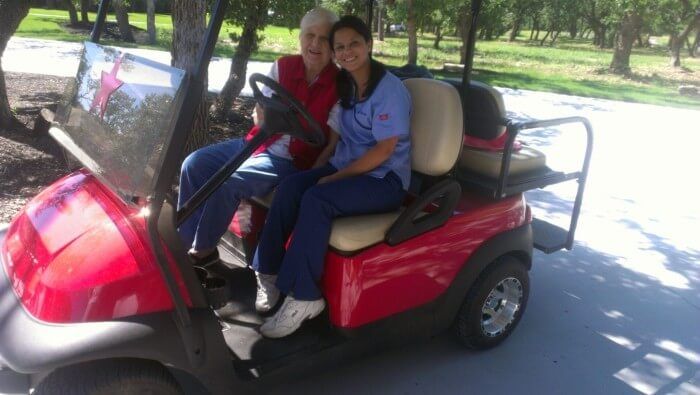 Two people in a red golf cart, smiling, on a paved path near trees. One wears scrubs.