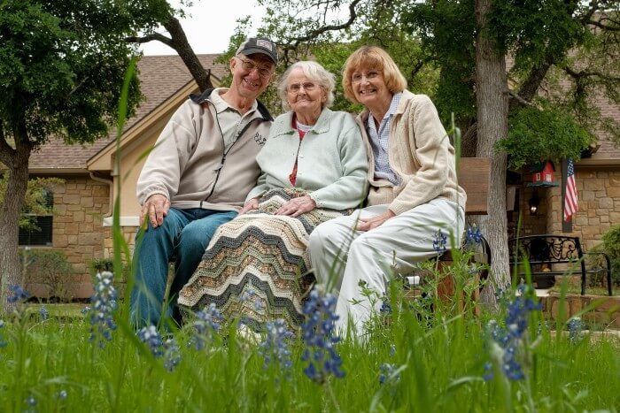 Family sits together on a bench in front of a stone house, blue flowers in foreground.