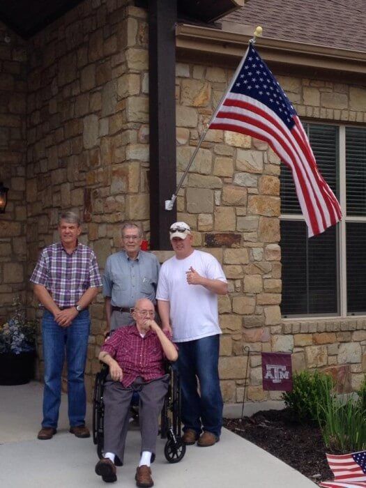 Four men posing outside a stone house with an American flag. One man sits in a wheelchair, others standing.