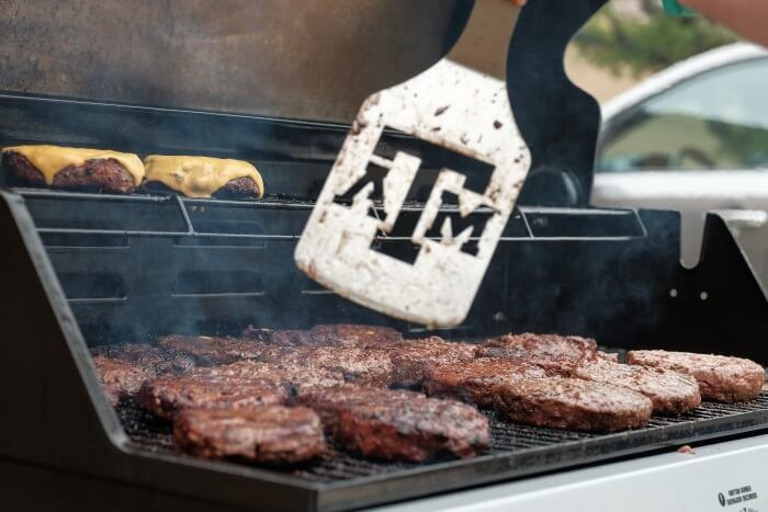 Burgers on a grill with a spatula showing the Texas A&M logo being used to flip them.