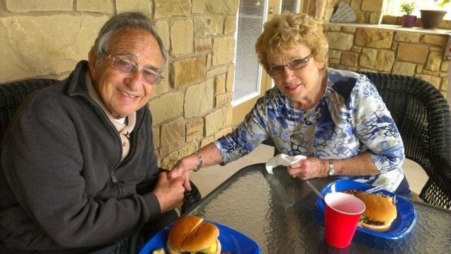 Elderly couple seated outdoors, smiling, holding hands, eating burgers, drinks on table.