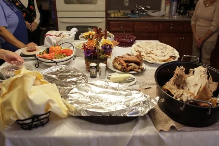 A table laden with Thanksgiving food: turkey, vegetables, rolls, and desserts. People are gathered.