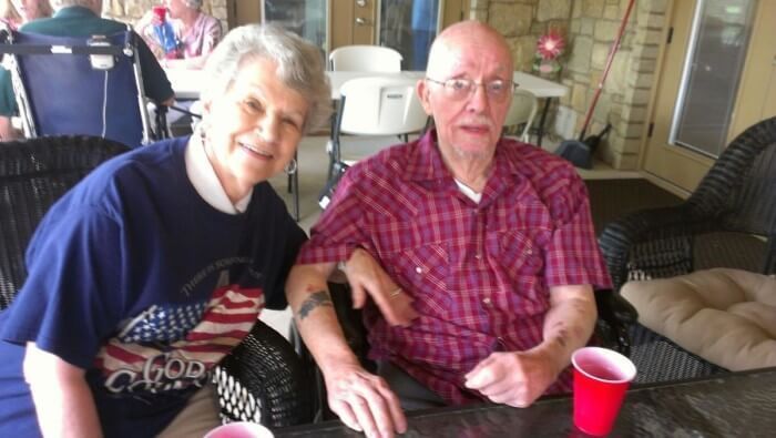 Elderly couple seated outdoors, smiling. Woman in blue shirt with American flag, man in red plaid shirt.