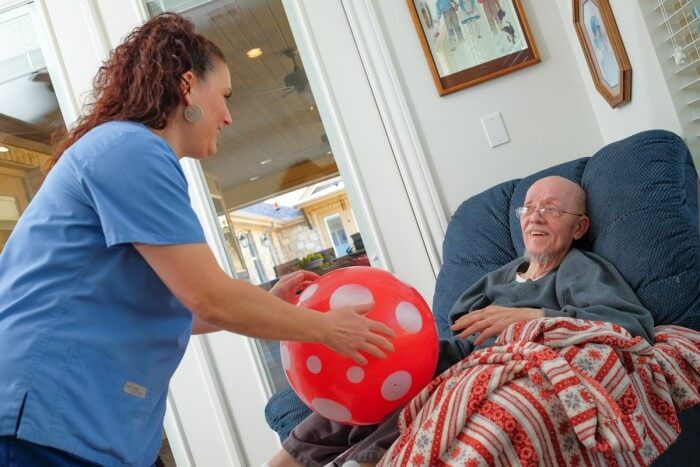 Caregiver offers a red and white polka dot ball to a person seated in a chair, inside a home.