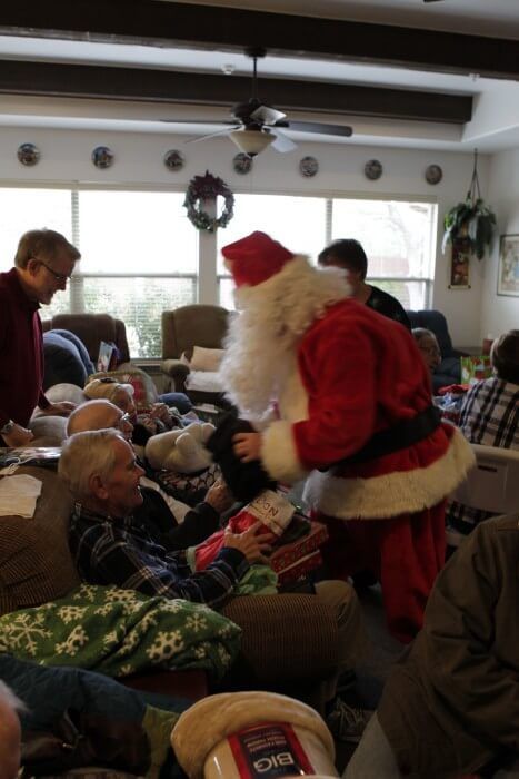 Santa hands a gift to a person sitting on a couch in a living room; others watch.