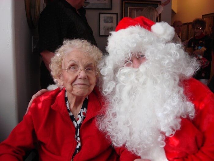Elderly woman in red jacket smiles next to Santa Claus.