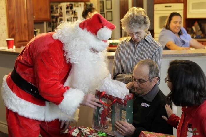 Santa Claus hands a wrapped gift to a man seated at a table, with a woman and child beside him.