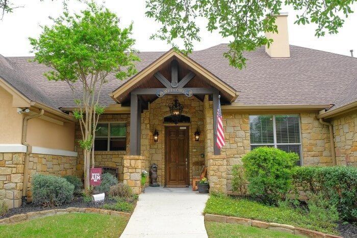 Tan brick home with a wooden front door under a brown portico. American flag hangs; landscaped front yard.